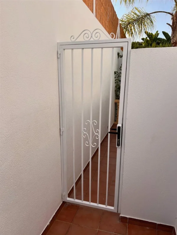 Puerta de reja de forja blanca con volutas en un pasillo exterior
